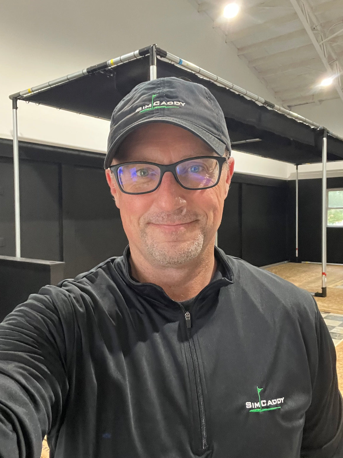A man wearing a black SimCaddy cap and a black zip-up shirt with the SimCaddy logo, taking a selfie inside an indoor simulator range with a black canopy visible in the background.